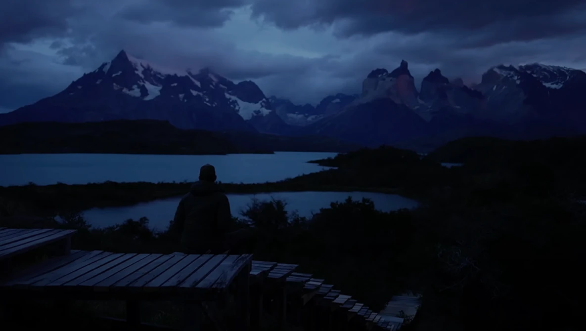 Silhouetted figure sitting on a wooden deck overlooking a mountain lake at dusk.