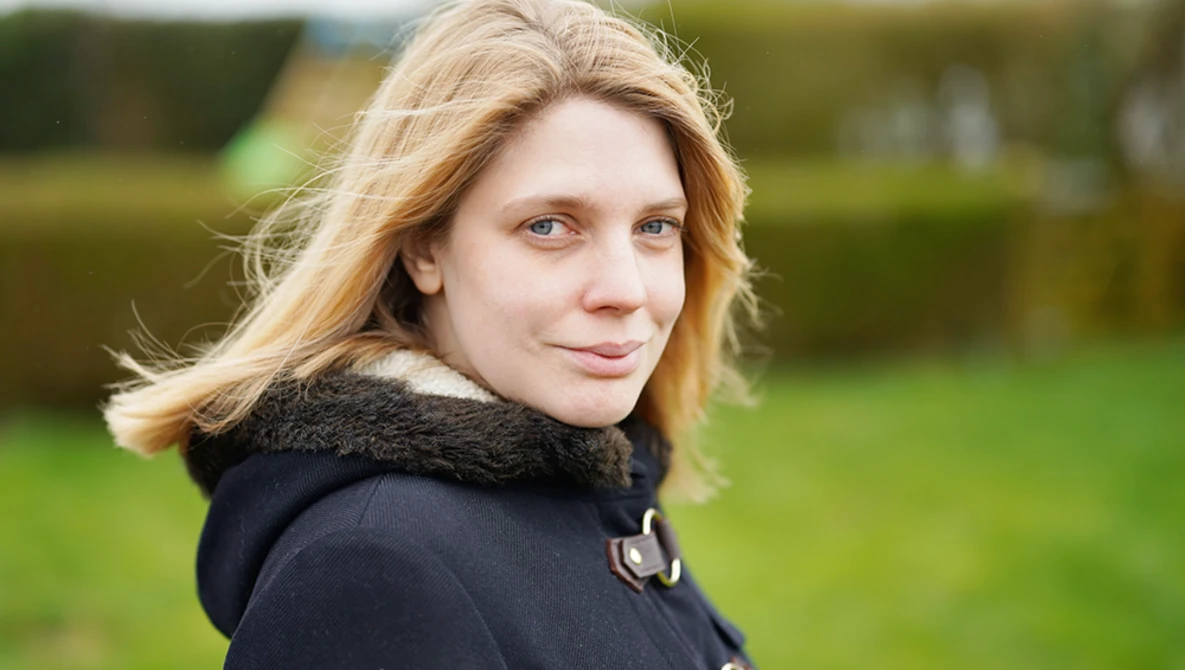 Woman with blonde hair looking over shoulder outdoors, wearing black jacket with fur collar.