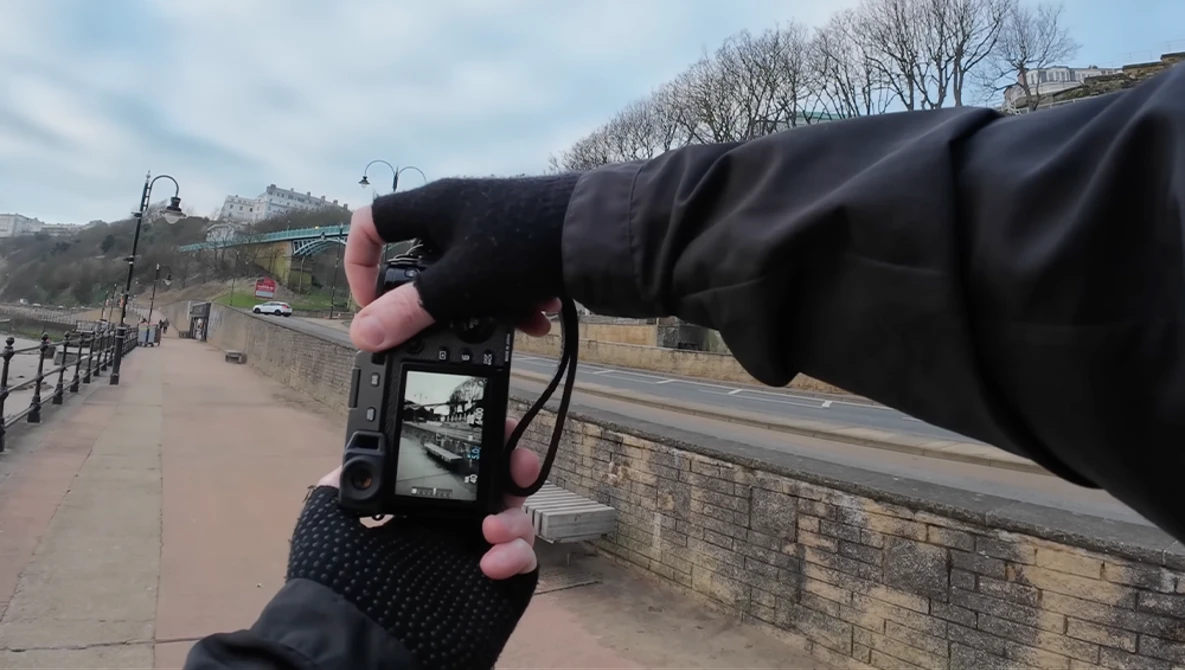Photographer's hands holding a smartphone to frame a riverside promenade with bare trees and stone walls.
