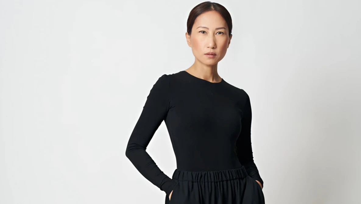 Woman in black long-sleeve top and gathered skirt standing against neutral background.