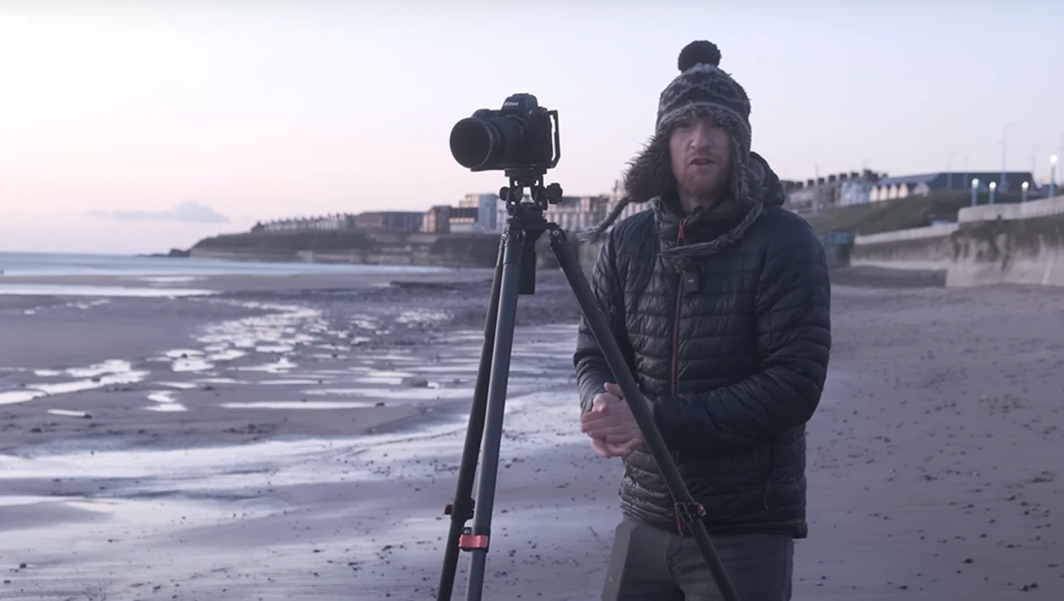 Photographer with camera on tripod on a beach at dusk, wearing winter clothing and beanie.