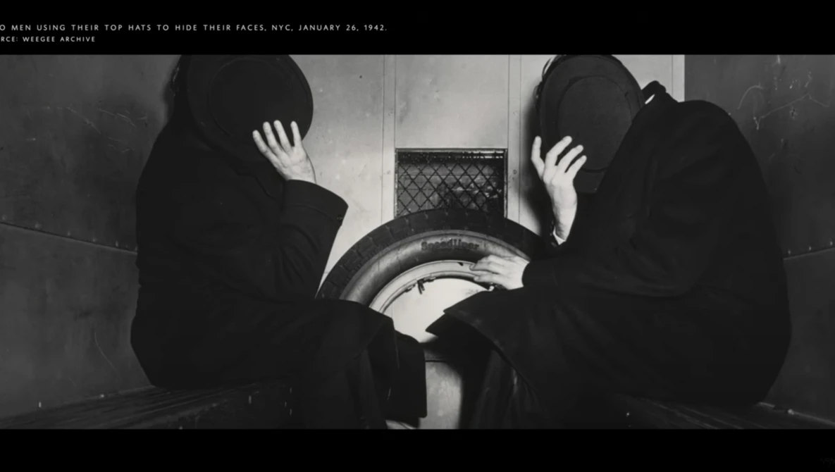 Two figures in dark clothing hide their faces with their hands while sitting near a large cylindrical metal object.