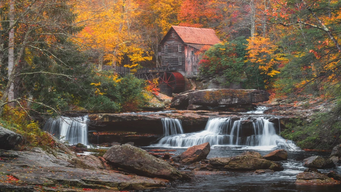 Historic gristmill with red roof beside cascading waterfall in autumn forest.