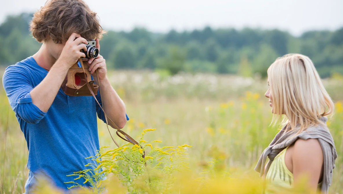 Young man in blue shirt photographing a woman in a natural outdoor field setting.