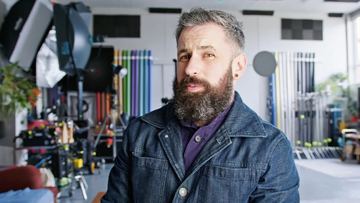 Bearded man in denim jacket inside a photography studio with organized equipment and lighting setups visible in the background.