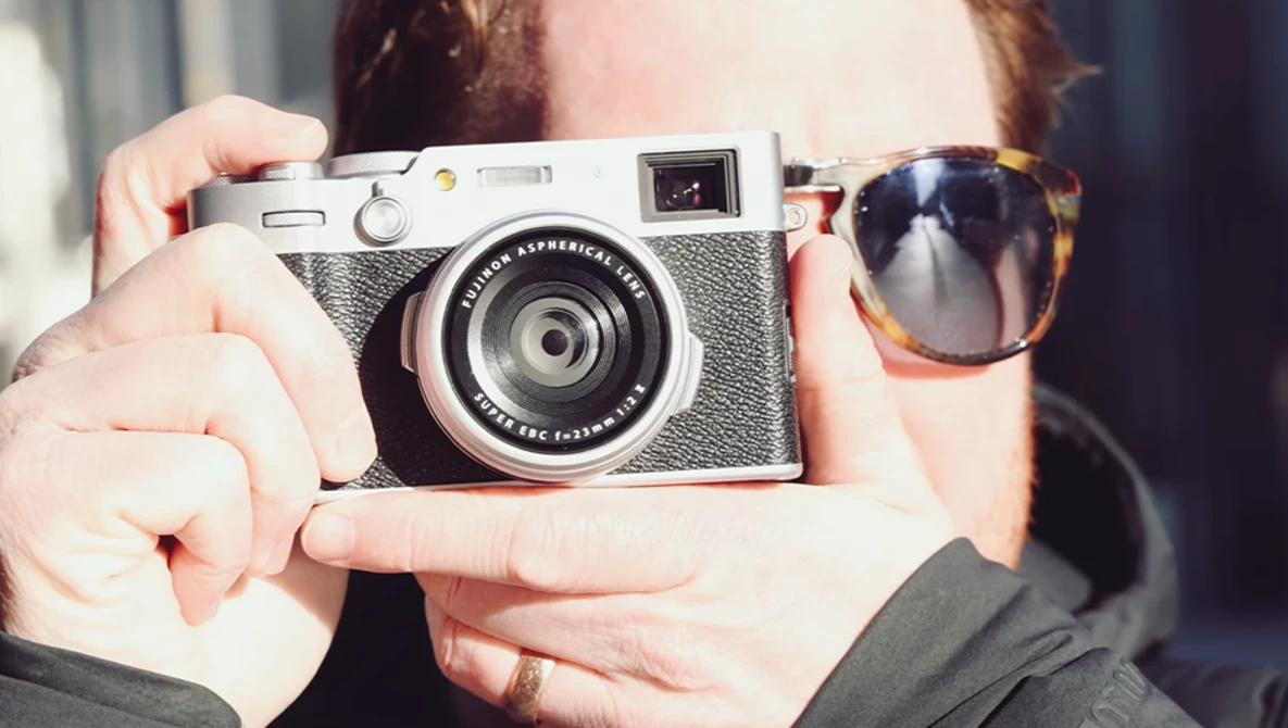 Person holding a rangefinder film camera with a fixed lens, wearing tortoiseshell sunglasses.
