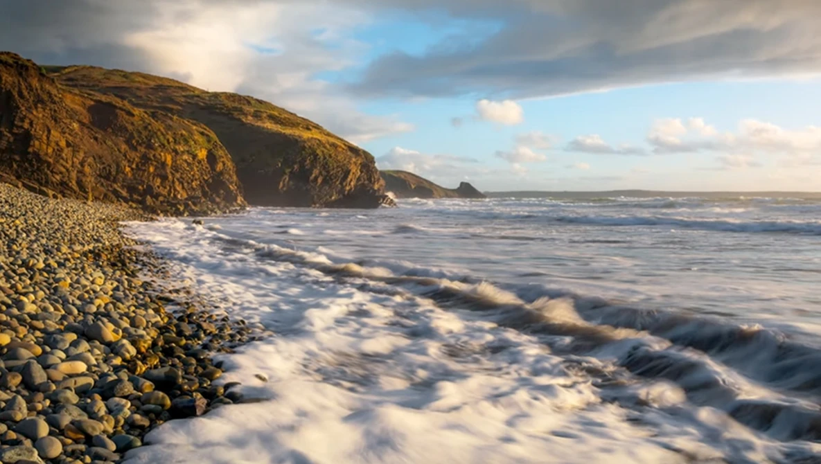 Rocky coastline with steep cliffs and turbulent waves rolling onto a pebble beach at golden hour.