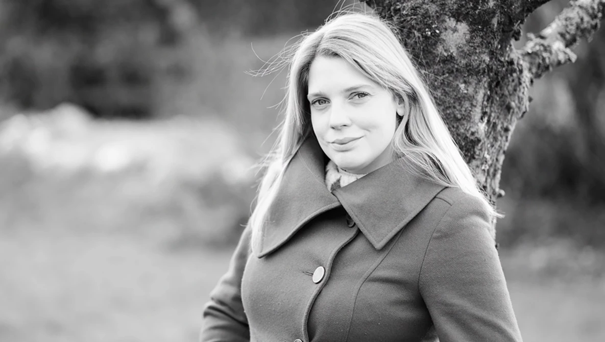 Woman with long blonde hair wearing a collared coat, positioned against a tree in an outdoor setting.