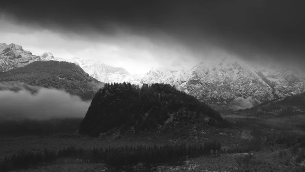 Snow-covered alpine peaks rise behind a forested ridge shrouded in mist under dark storm clouds.