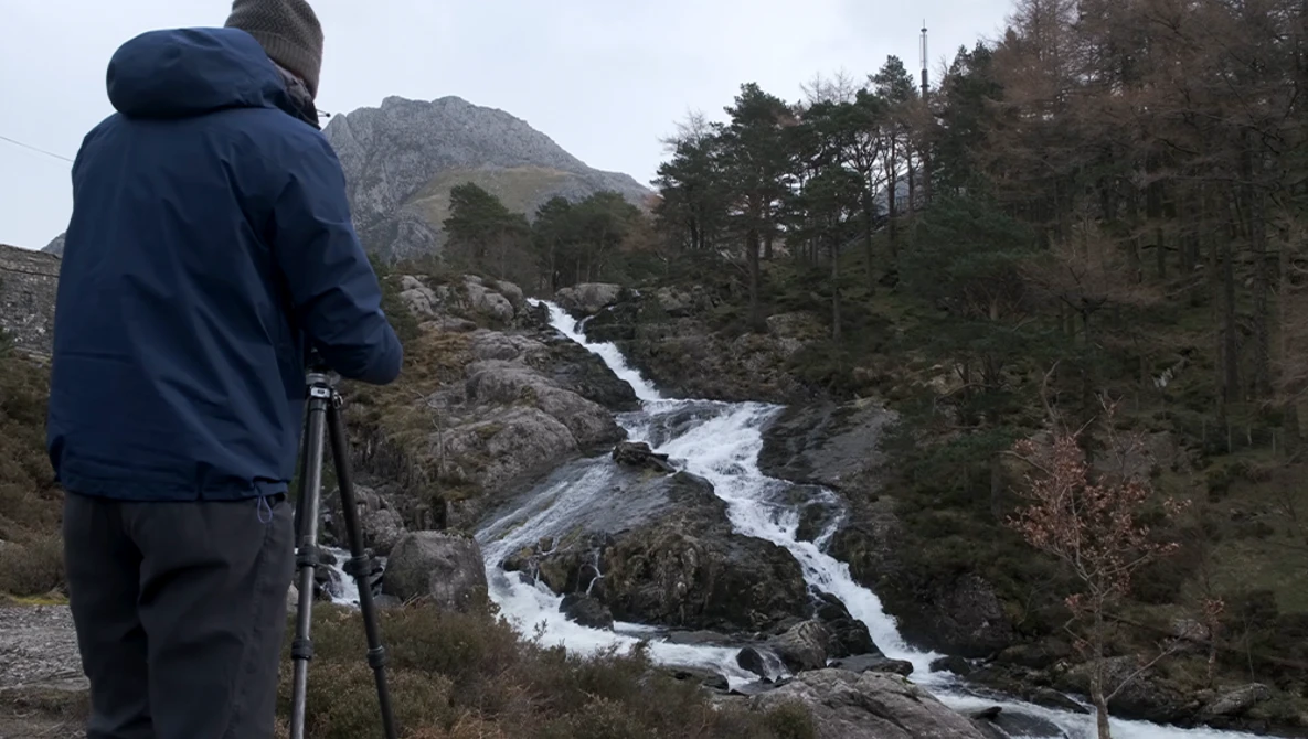 Photographer on tripod capturing a cascading mountain stream surrounded by evergreen forest and rocky terrain.