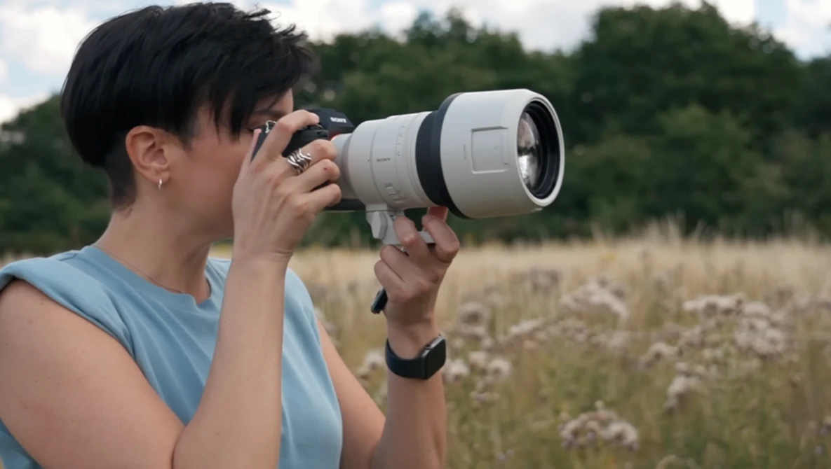 Photographer holding a telephoto lens camera up to her eye in an open field with wildflowers and trees in the background.