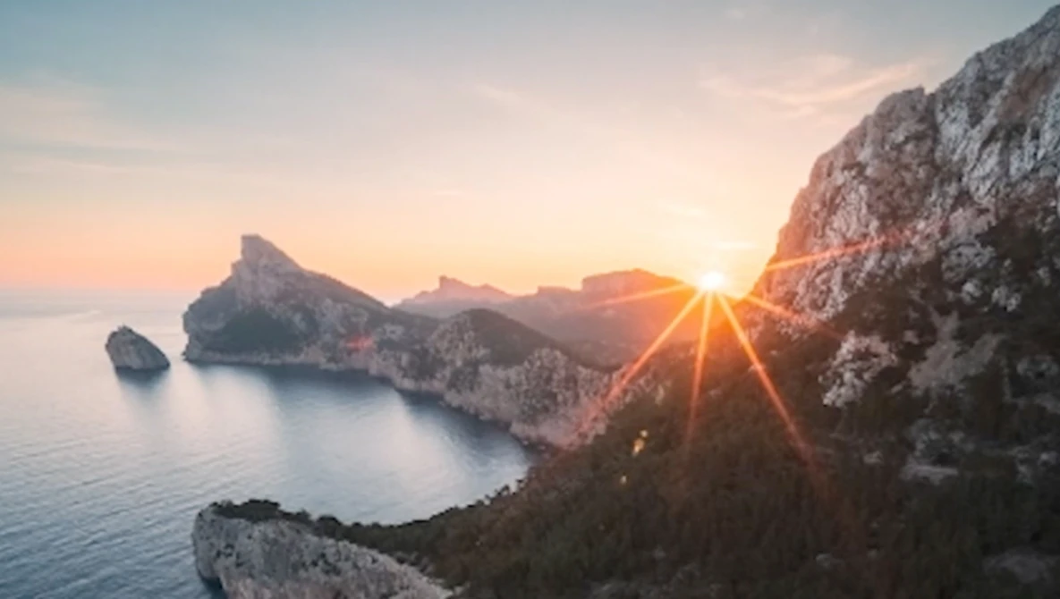 Sun bursting through rocky cliffs above a calm Mediterranean cove at sunrise.