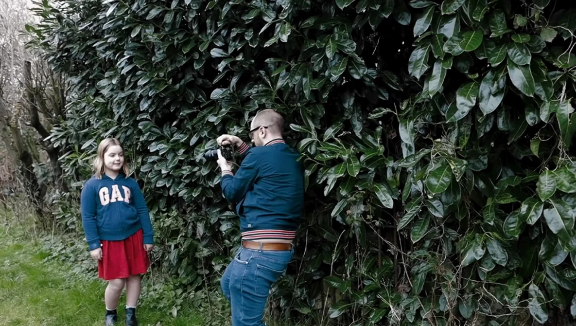 Photographer taking a portrait of a young girl standing against dense ivy-covered foliage.
