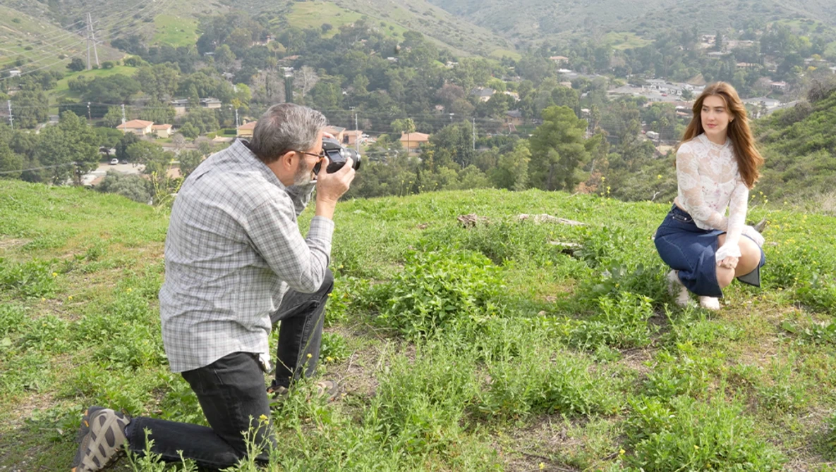Photographer crouching while shooting a model posing in a grassy hillside field with forested mountains in the background.