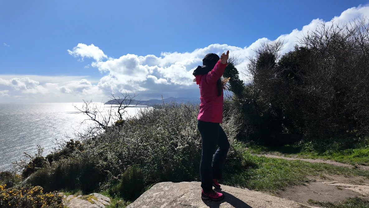 Person in magenta jacket photographing a coastal landscape with binoculars or telephoto lens.