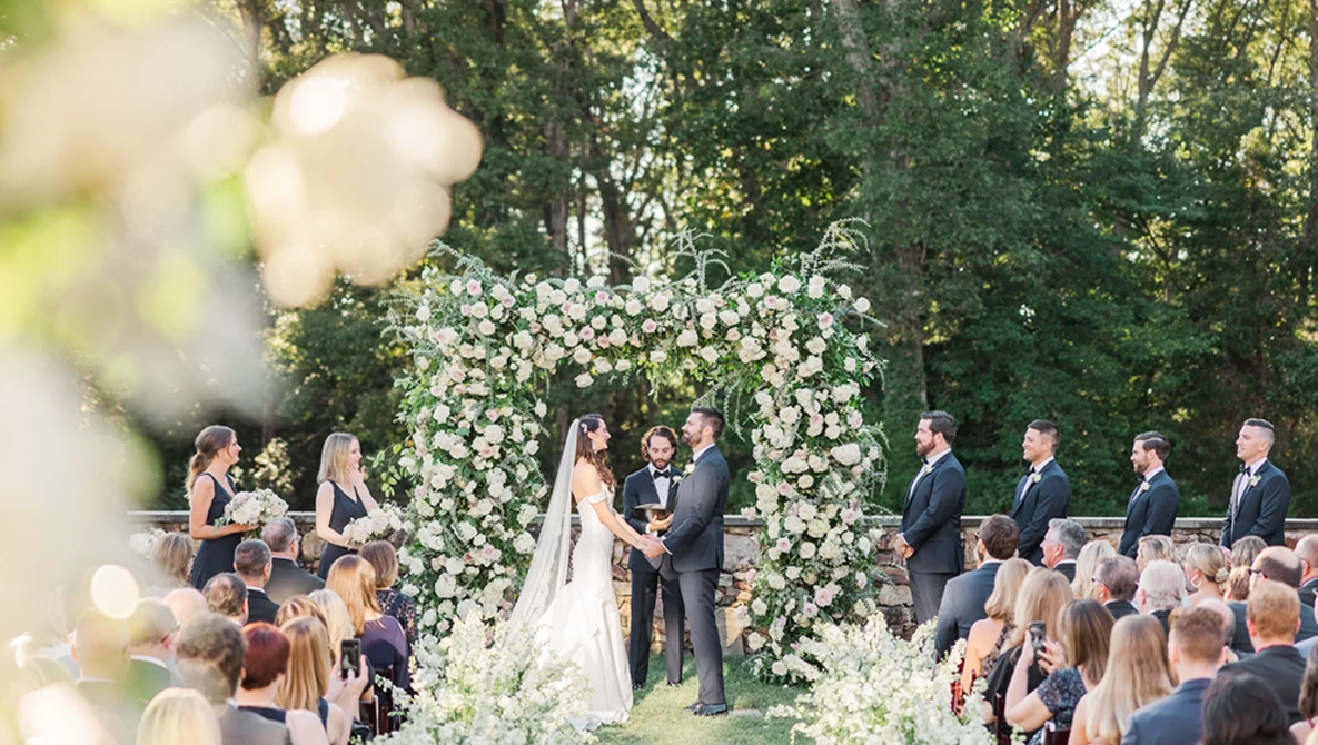 Bride and groom exchanging vows beneath an elaborate floral arch at an outdoor wedding ceremony.