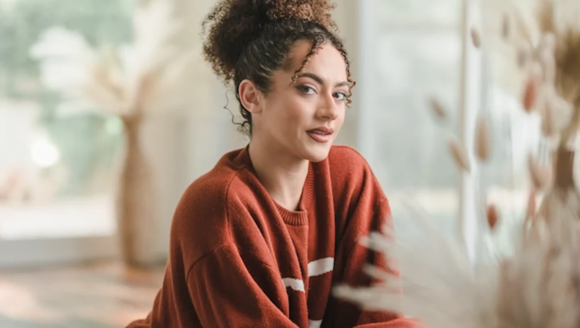 Woman in rust-colored sweater gazing to the side near a sunlit window.