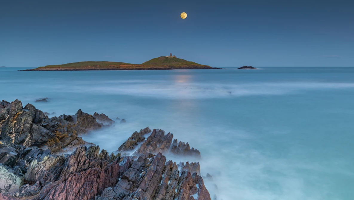 Rocky coastline with smooth water and distant island under clear sky with moon.
