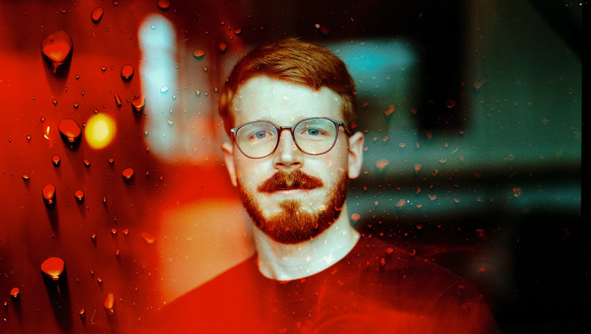 Man wearing glasses and red shirt photographed through rain-covered glass with warm and cool color cast.