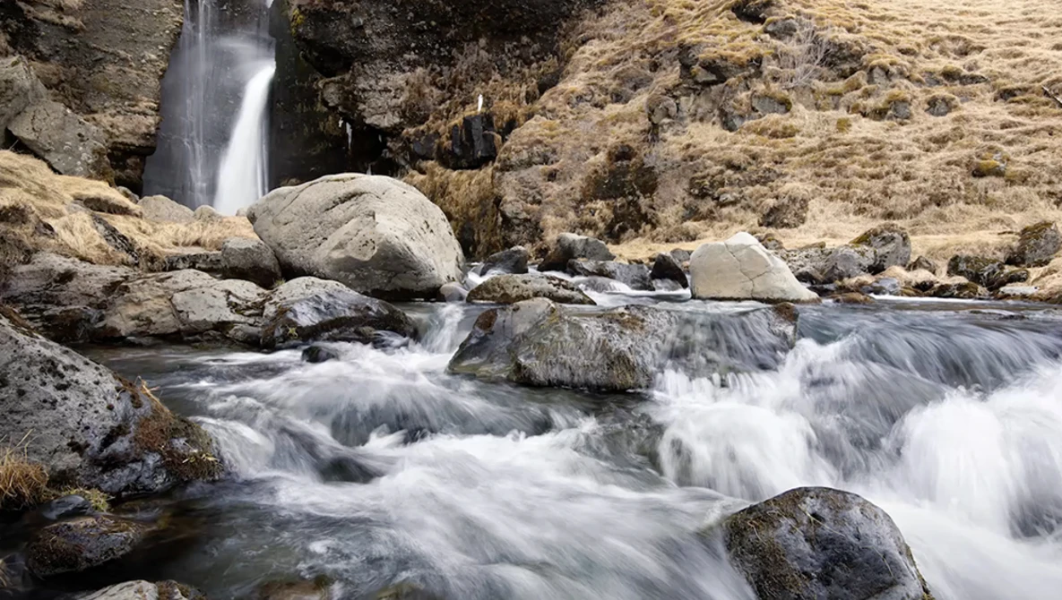 Mountain stream with cascading white water flowing around moss-covered rocks and boulders, with a waterfall visible in the background.