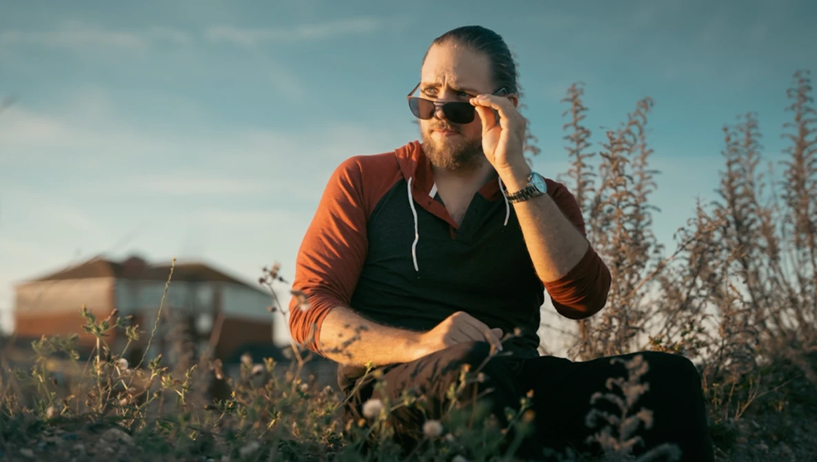Man in orange shirt and sunglasses crouching in a field during golden hour light.