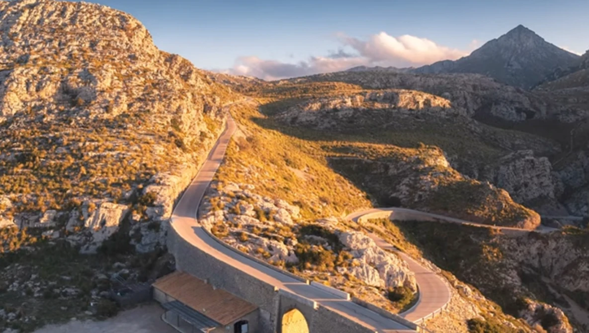 Winding mountain road cutting through golden-lit rocky peaks at sunset with a distant volcanic cone.