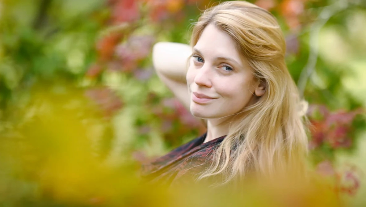Young woman with long blonde hair looking over her shoulder in an outdoor garden setting.