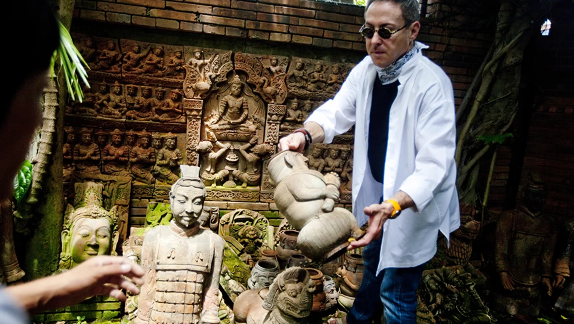 Man in light blue shirt gesturing toward stone sculptures in an overgrown courtyard with carved relief panels.