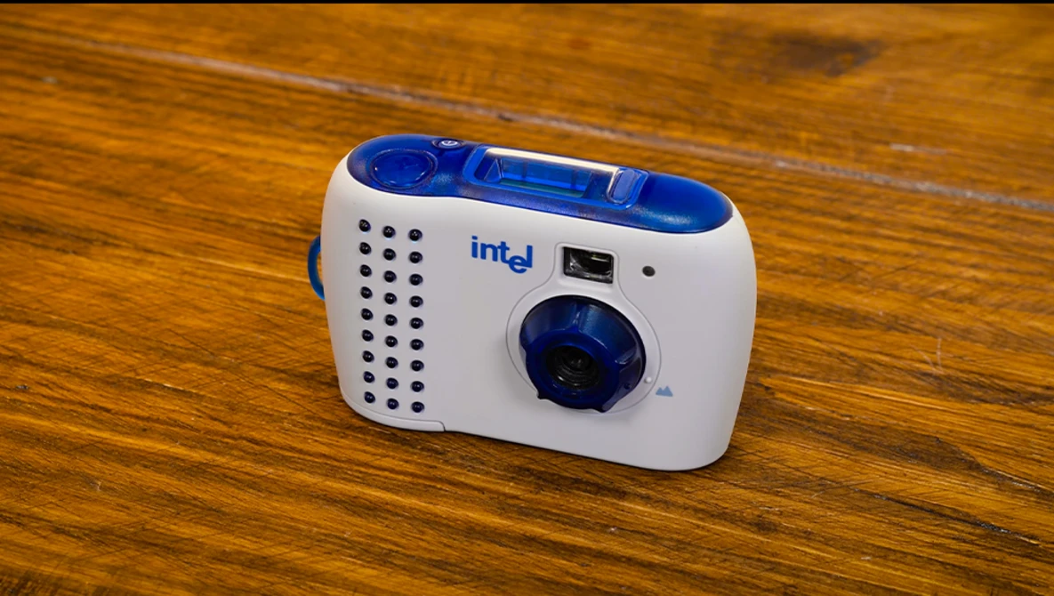 Intel-branded compact point-and-shoot camera with white body, blue accents, and perforated speaker grille on wooden surface.