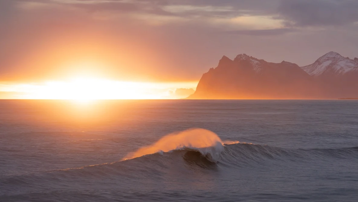 Ocean wave with golden spray backlit by sunset, snow-capped mountain silhouetted on horizon.