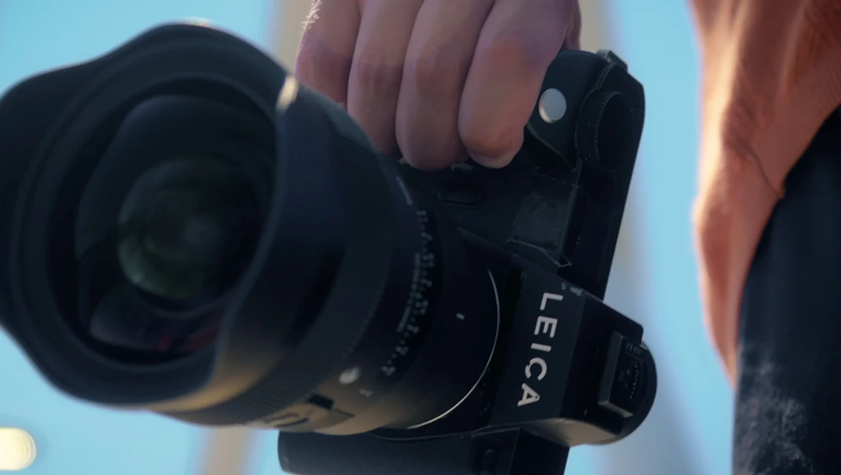 Photographer holding a mirrorless camera with a wide-angle lens against a blue sky background.