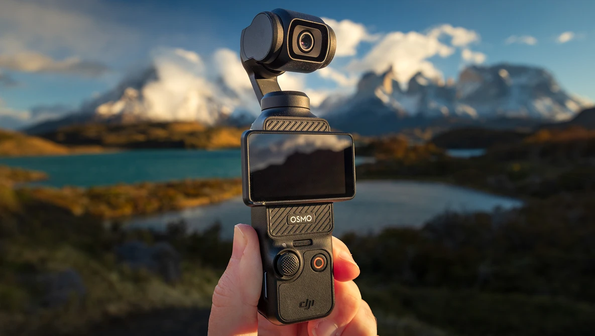DJI Osmo Pocket gimbal camera held in hand against snowy mountain peaks and turquoise lake landscape.