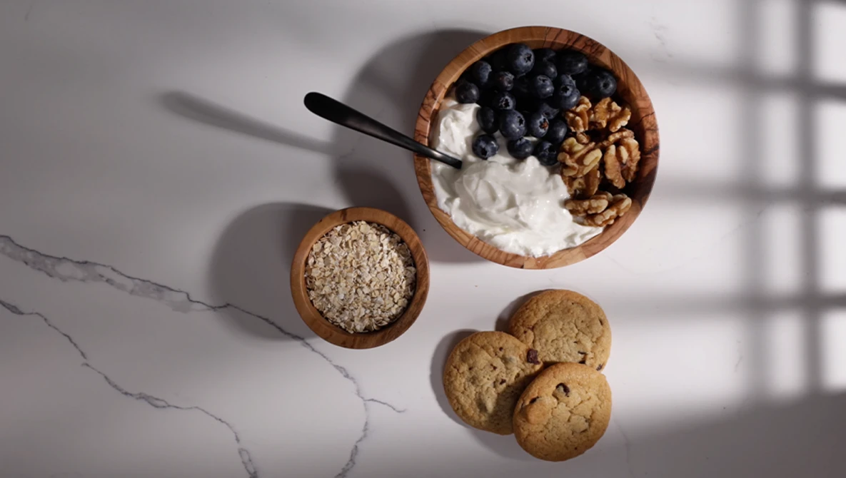 Overhead flat lay of breakfast ingredients in wooden bowls: yogurt with blueberries and granola, oats, and cookies on marble surface.