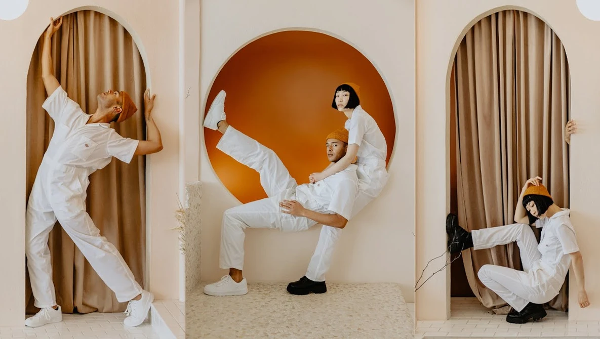 Three people in white clothing posing dynamically in arched doorways with tan curtains and an orange circular backdrop.