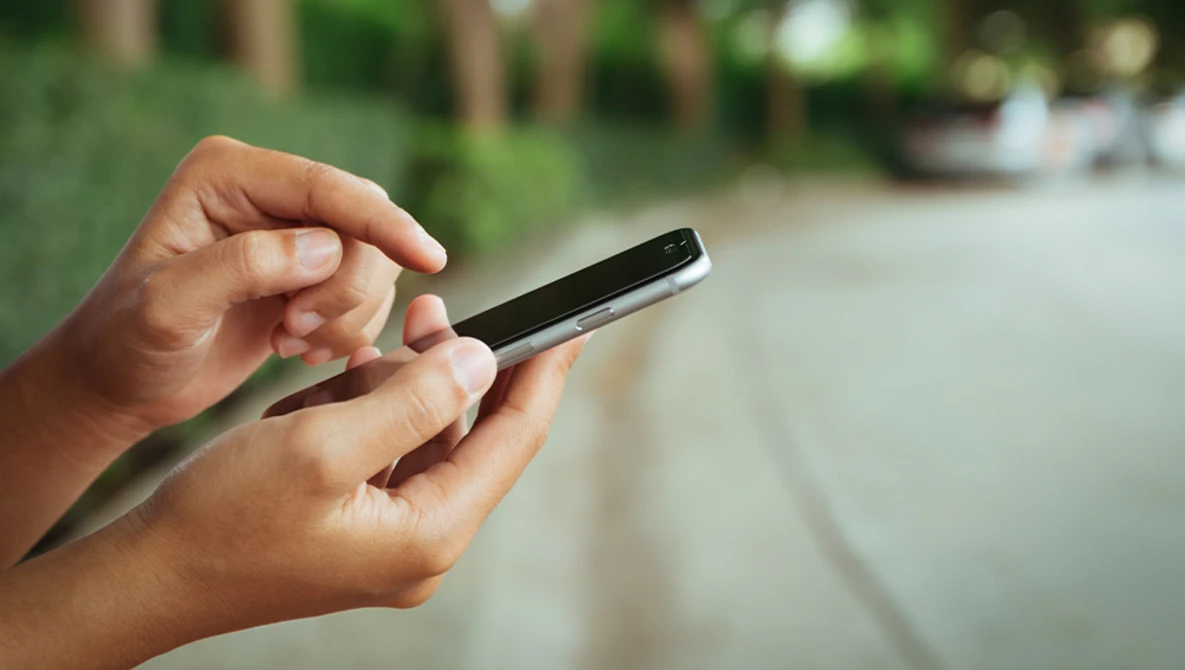 Hands holding a smartphone outdoors with blurred green foliage in the background.