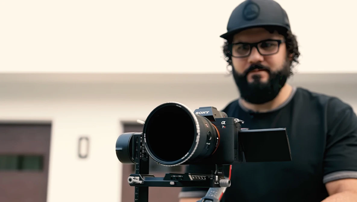 Photographer with a large telephoto lens mounted on a tripod head, wearing a baseball cap and glasses.