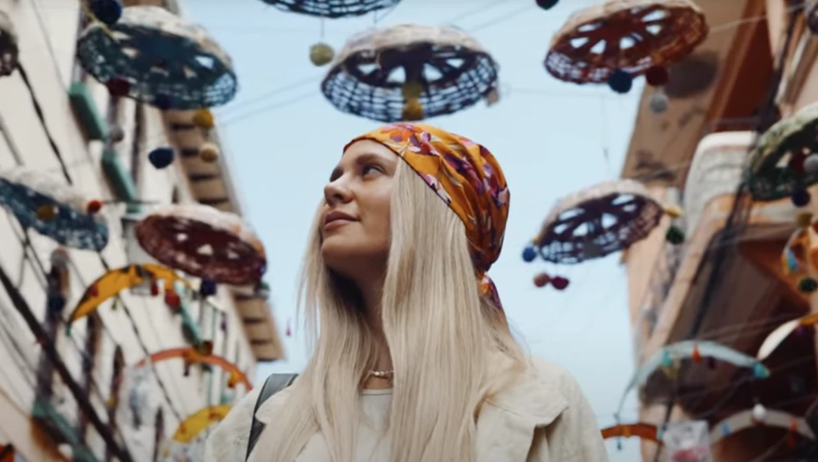 Woman with long platinum blonde hair and gold headwrap gazing upward surrounded by hanging decorative UFO sculptures.