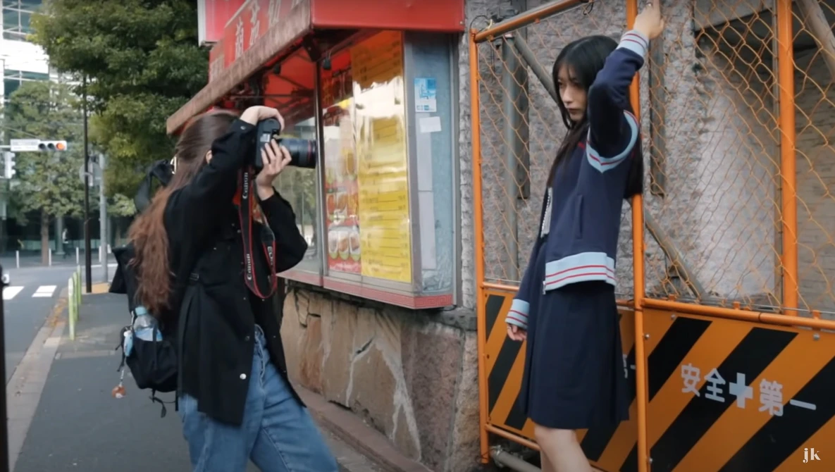 Two students in school uniforms posing playfully against urban storefronts and metal structures.