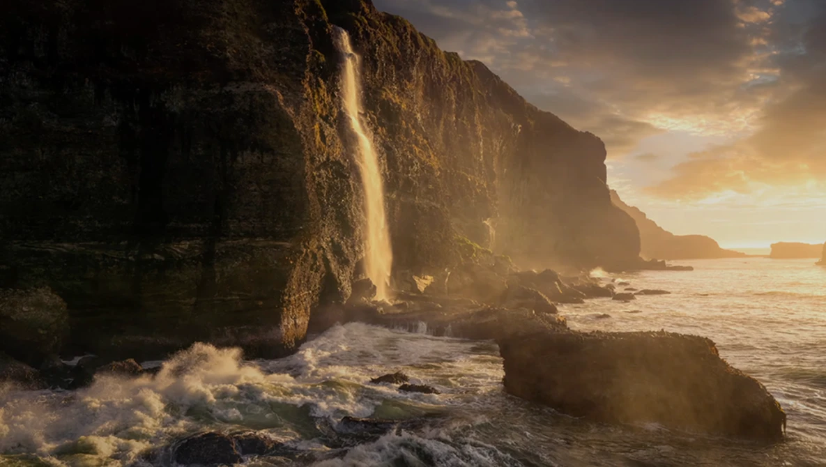 Waterfall cascading down a steep coastal cliff face during golden hour, with waves crashing on rocks below.