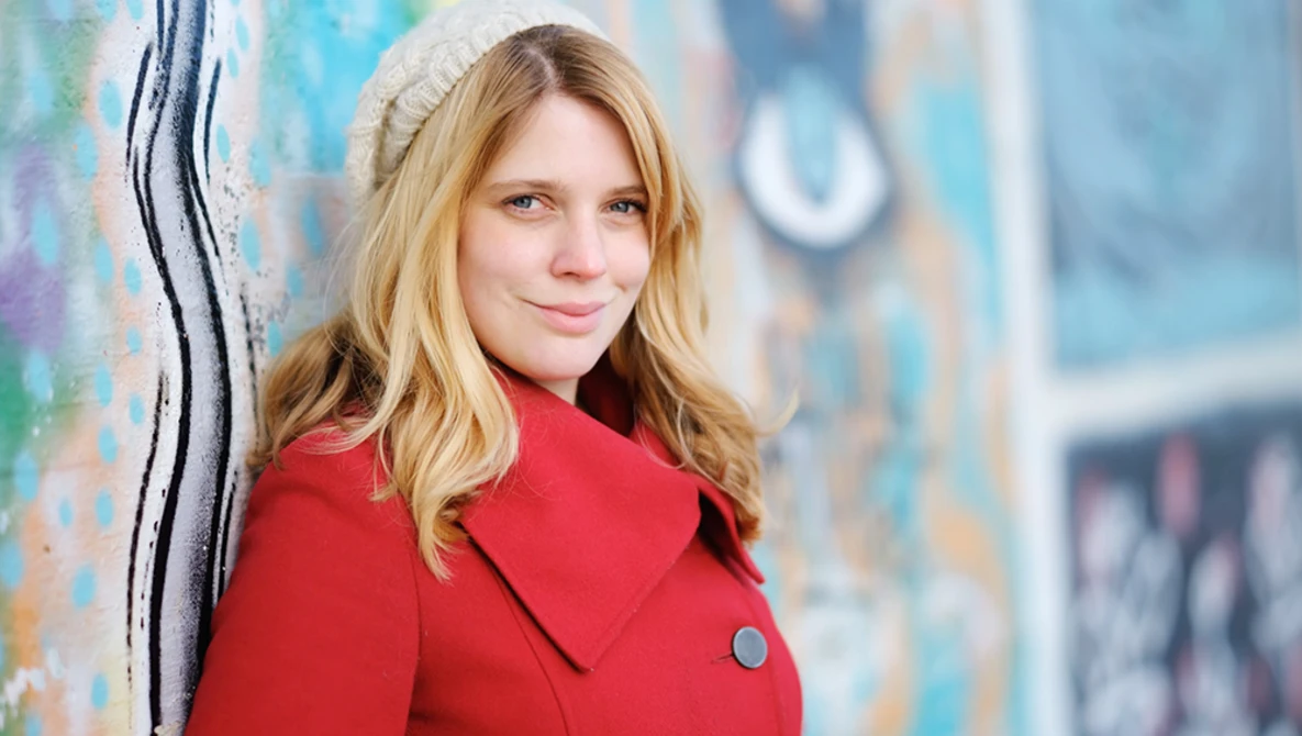 Portrait of a young woman with blonde hair wearing a red coat against a colorful textured wall.