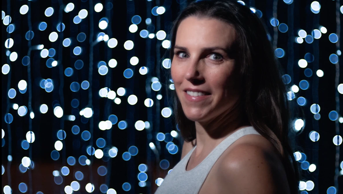 Woman with long dark hair smiling at camera against bokeh light background.