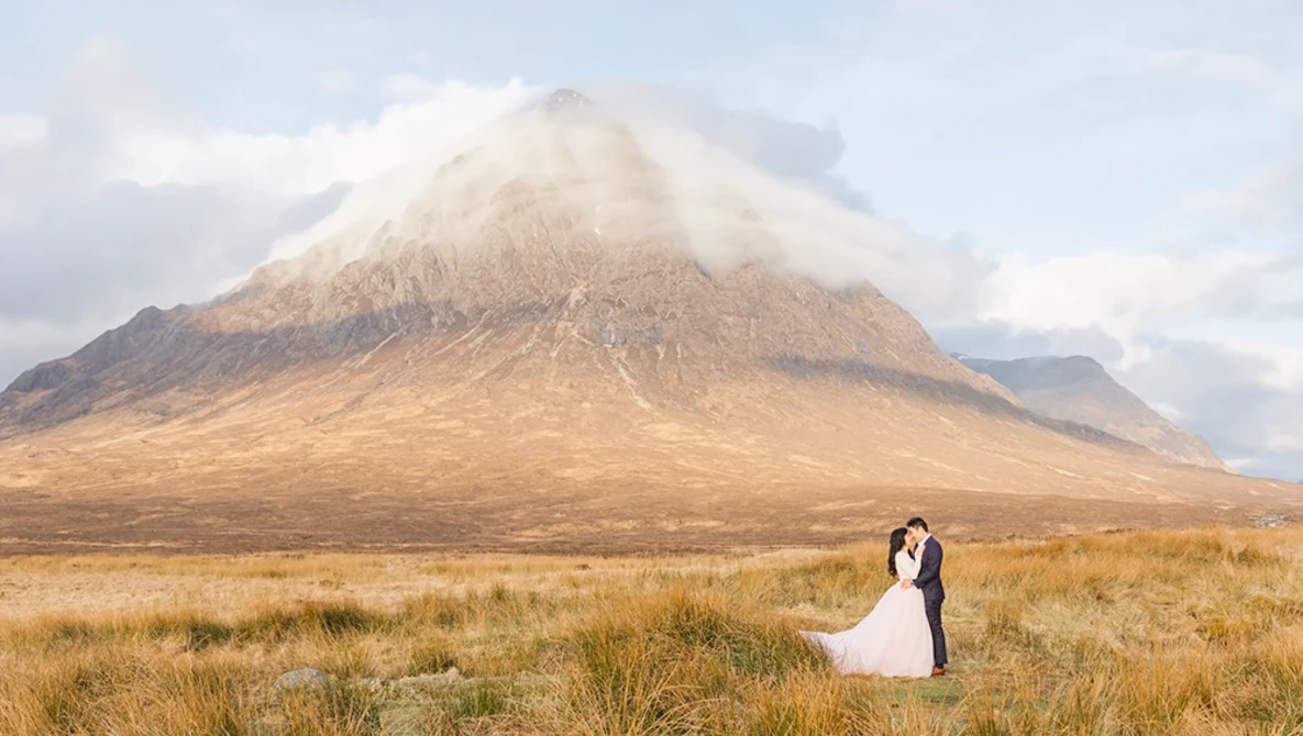 Bride and groom embracing in a vast golden grassland with a snow-capped volcanic mountain in the background.