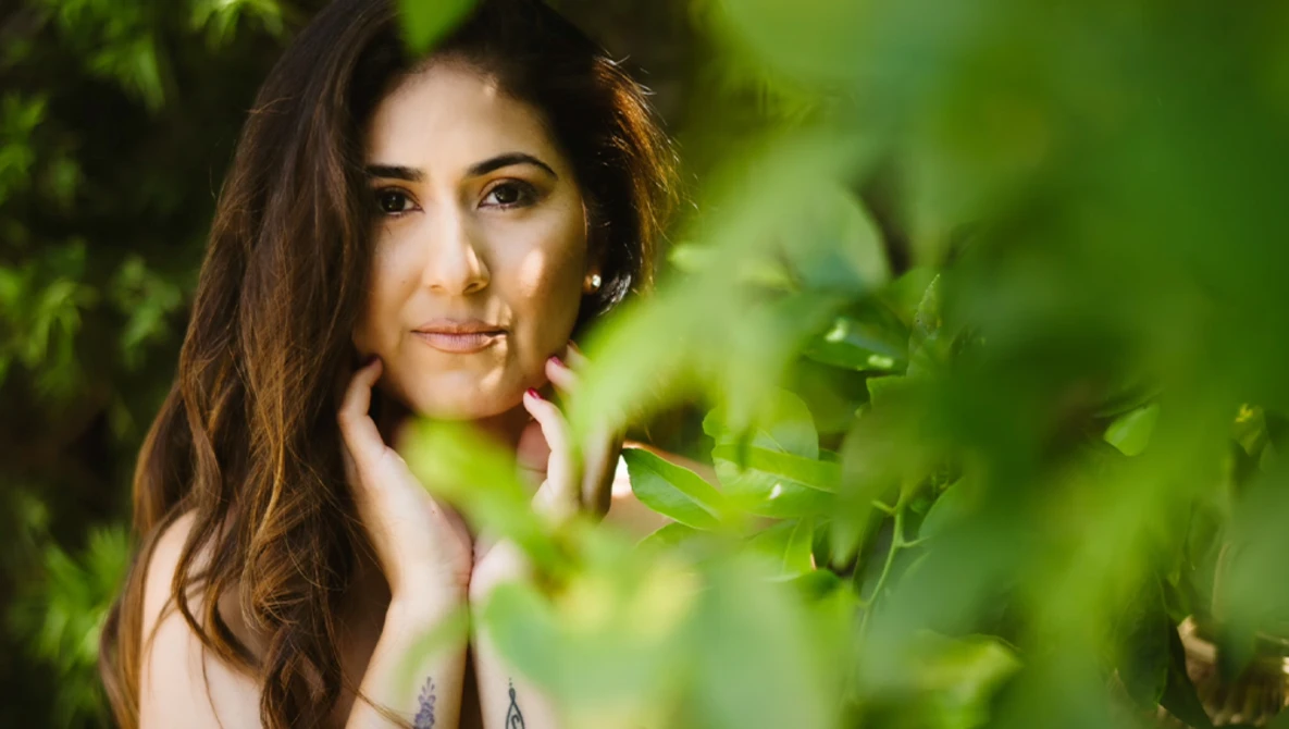 Woman with long brown hair posing among green foliage with soft natural lighting.
