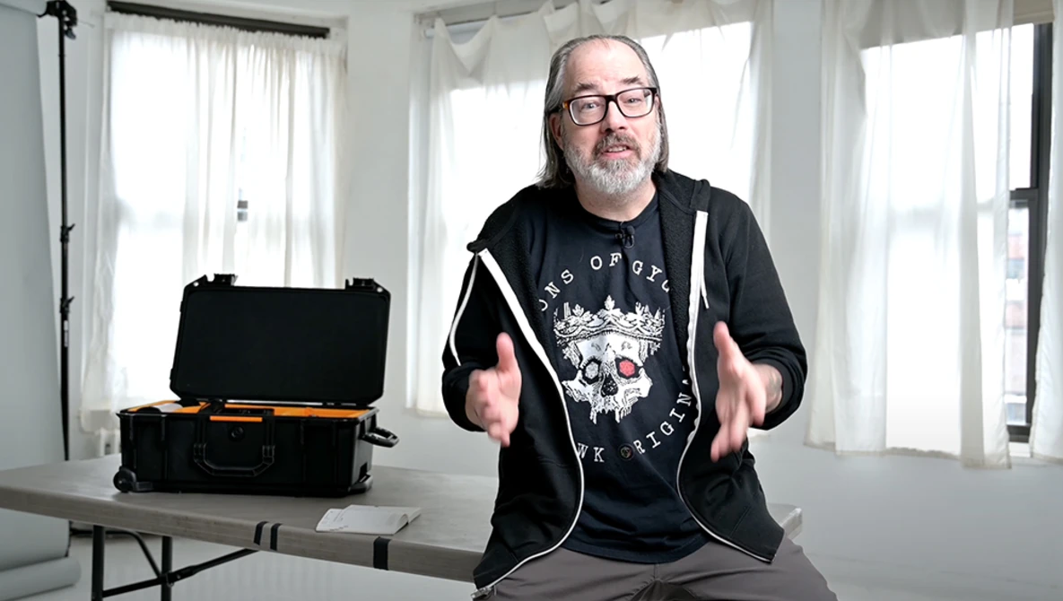 Man in glasses and black hoodie gestures while speaking in bright studio with equipment case on table.