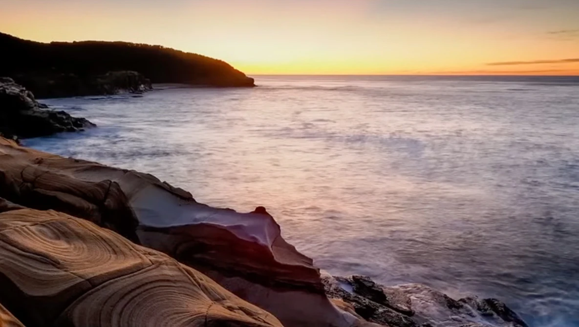 Rocky coastline at sunset with layered sandstone formations and calm ocean waters.