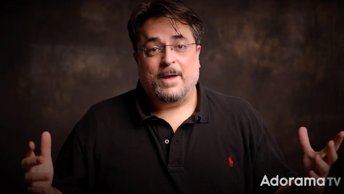 Man in black polo shirt gesturing while speaking against a dark textured background.