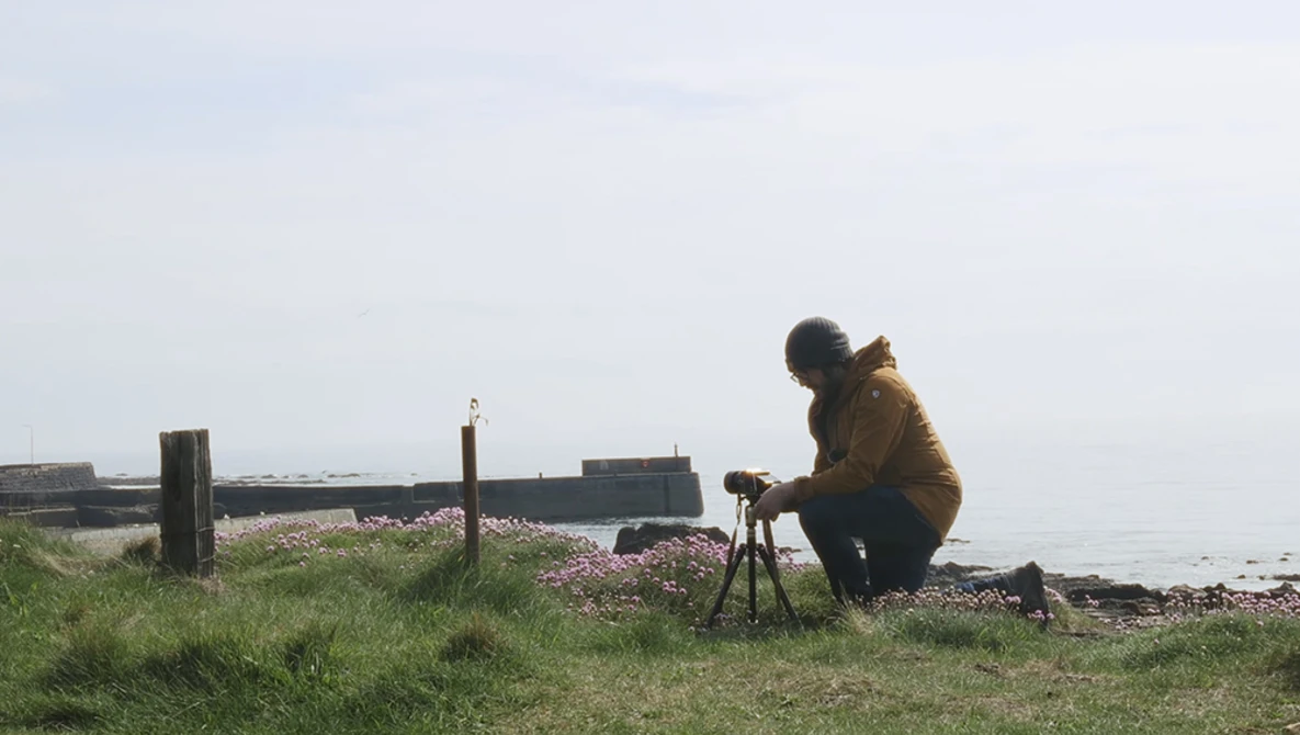 Photographer crouching with tripod-mounted camera on a pebbly beach overlooking water.