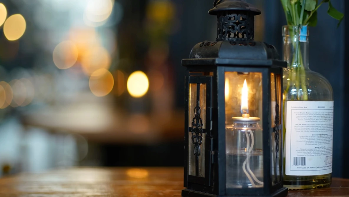 Black metal lantern with lit candle next to a wine bottle on wooden surface with bokeh lights in background.