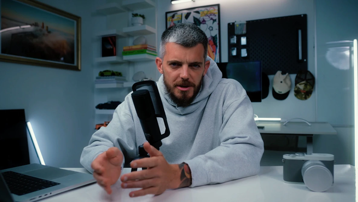 Man in gray hoodie demonstrating a handheld camera or recording device at a desk.