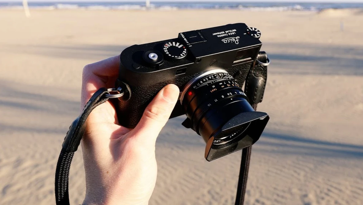 Hand holding a rangefinder camera with attached lens against a beach background.